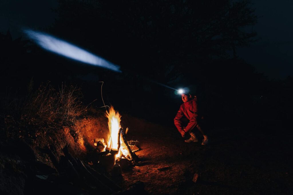 Man with headlamp sitting by campfire at night, surrounded by nature.