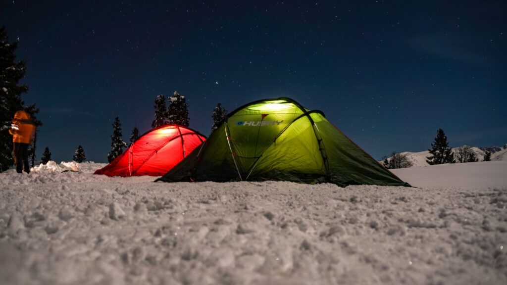 Two tents illuminated at night, set on a snowy landscape beneath a clear starry sky.