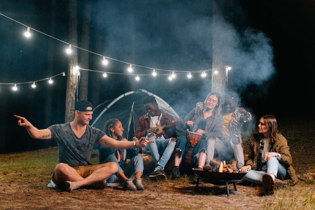 A group of friends relaxes by a campfire in the forest, playing music and talking.