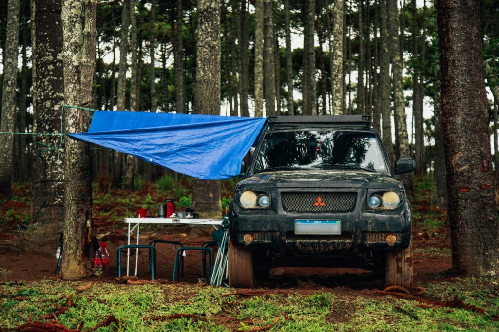 A rugged off-road vehicle parked in a forest clearing, set up for a camp with tarp and table.