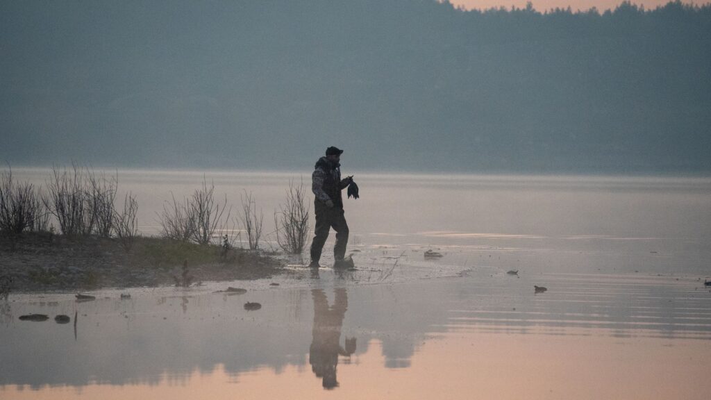A lone hunter walks along a foggy lakeside during sunrise, capturing the serene beauty of the morning.