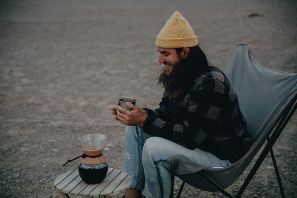 A relaxed man in a checkered coat with a yellow beanie enjoys coffee outdoors, embodying a rustic lifestyle.