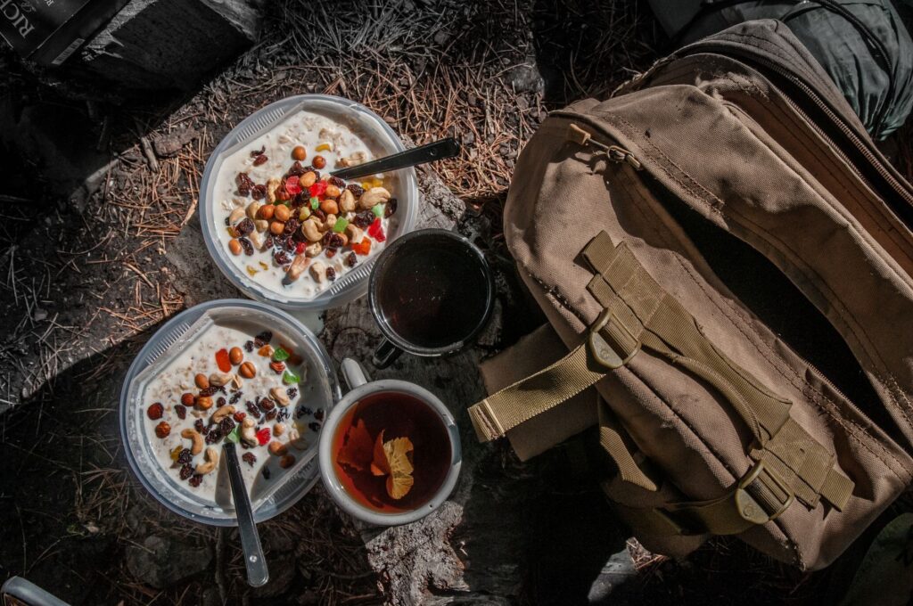 Top view of a rustic outdoor meal setup with fruit and nut bowls, tea, and backpack in a forest setting.
