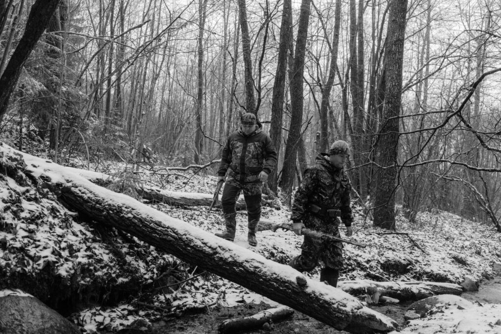 Two hunters in camouflage walking through a snowy forest, holding rifles. A monochrome winter scene.