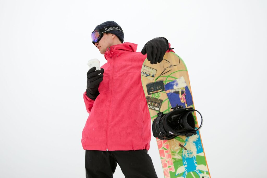 Man in winter clothing holds a snowboard and coffee, embodying a snowy adventure vibe.
