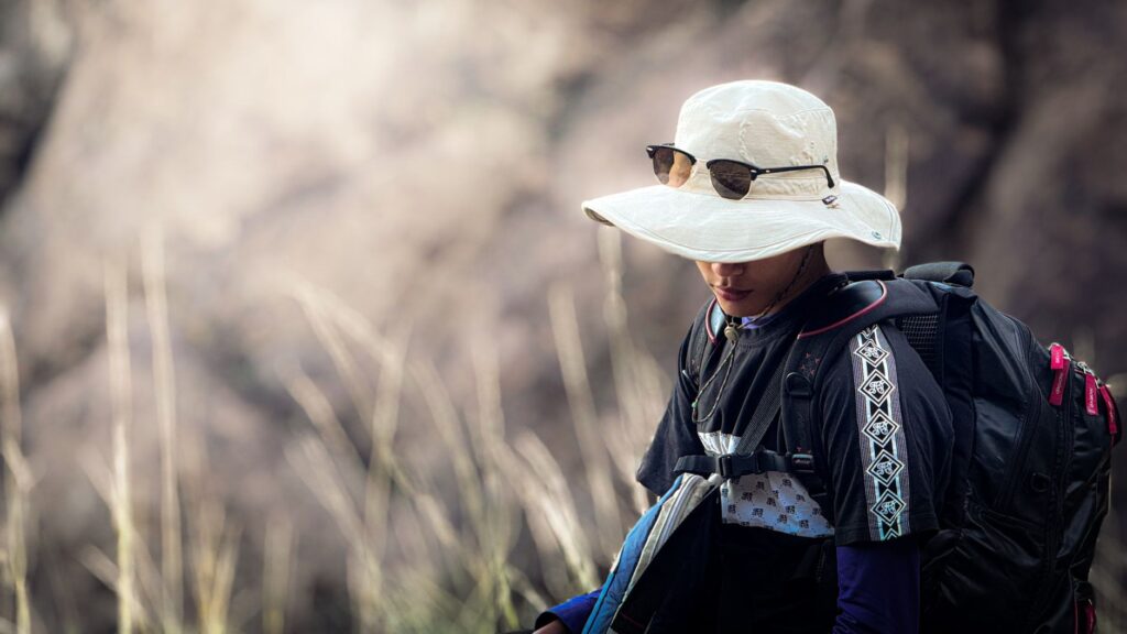 A young man wearing a sun hat and sunglasses hikes outdoors with a backpack, surrounded by natural scenery.
